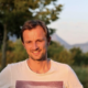 Smiling man with short brown hair wearing a white T-shirt stands outdoors with a blurred background of trees and mountains in soft, warm light.
